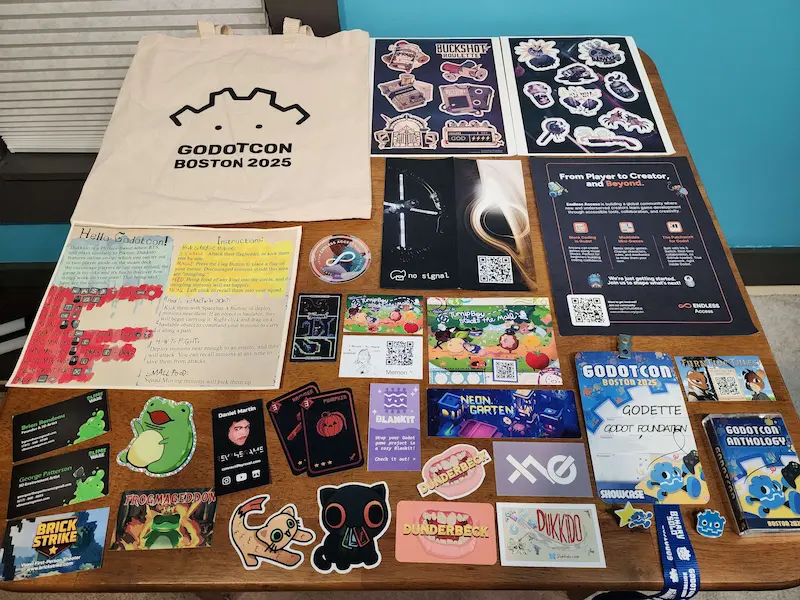 A table of goods collected at GodotCon. Godot branded tote, Buckshot Roulette sticker packs, cards and stickers from the showcase games, badge, pins, and the GodotCon Anthology Mixtape