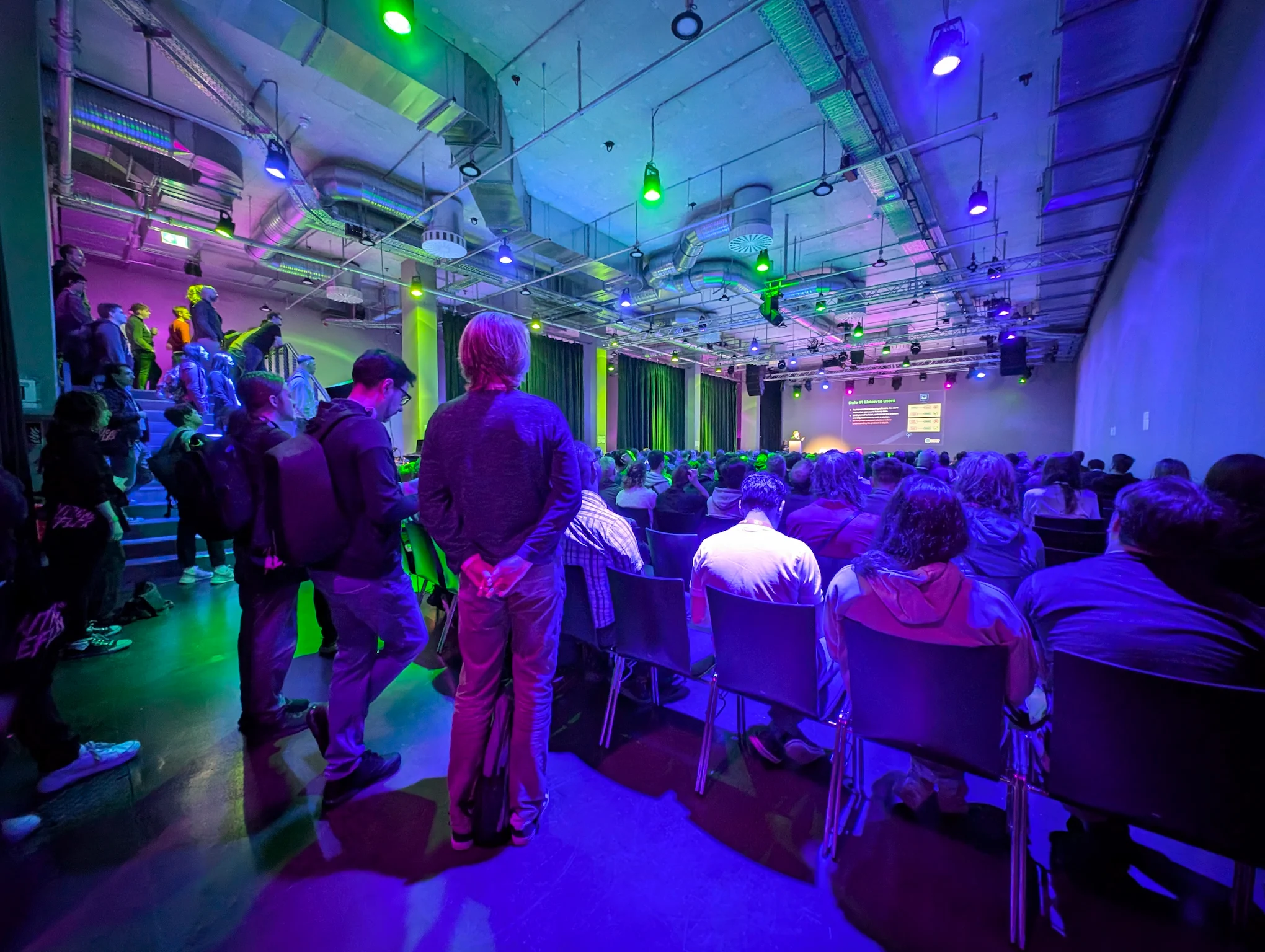 View of the stage, with people standing behind full rows of chairs.